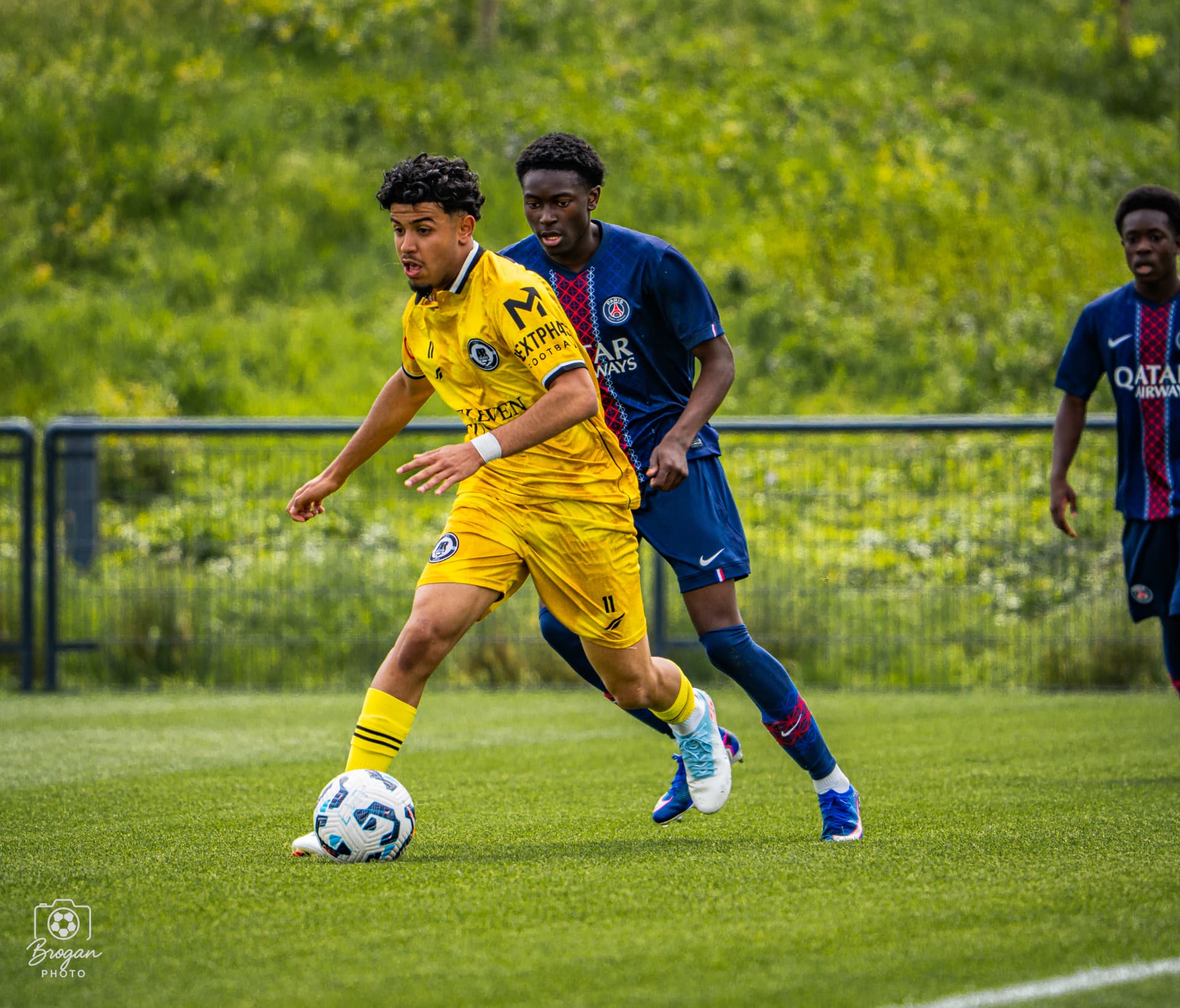 A Next Phase Football player driving past a Paris Saint-Germain defender during a competitive fixture.